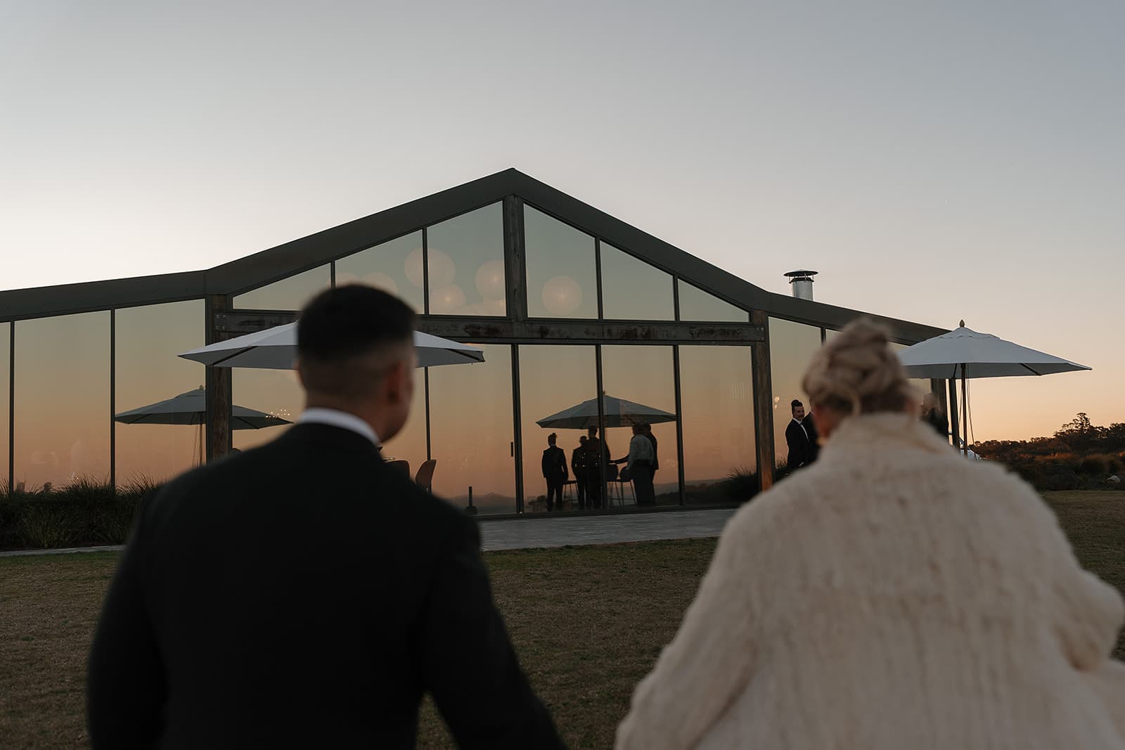 The couple walking back across the lawn to the Pavilion at dusk, its glass glowing amber with guests inside and umbrellas on the terrace