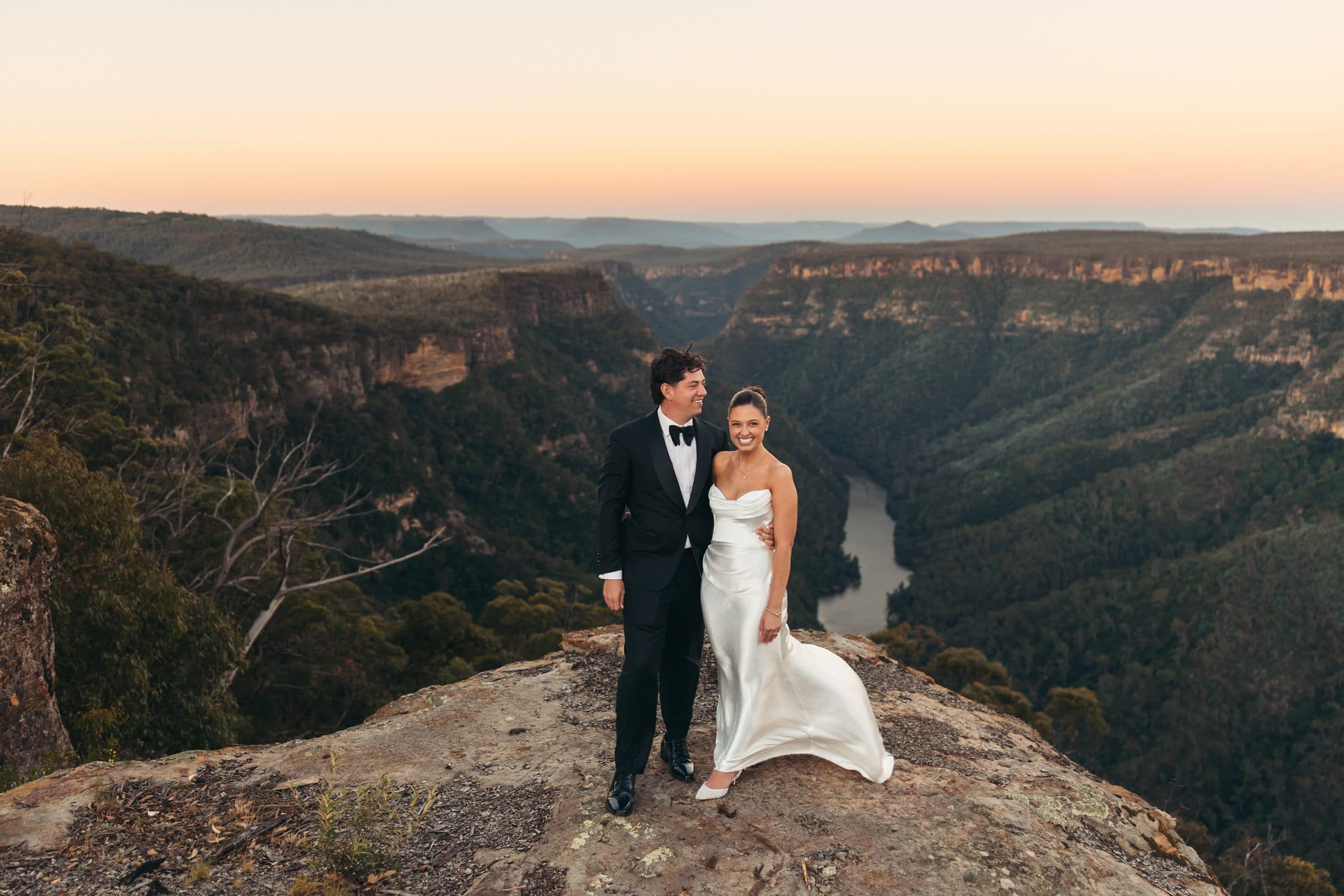 A couple standing on a sandstone ledge at sunset, high above the Shoalhaven River winding through the gorge