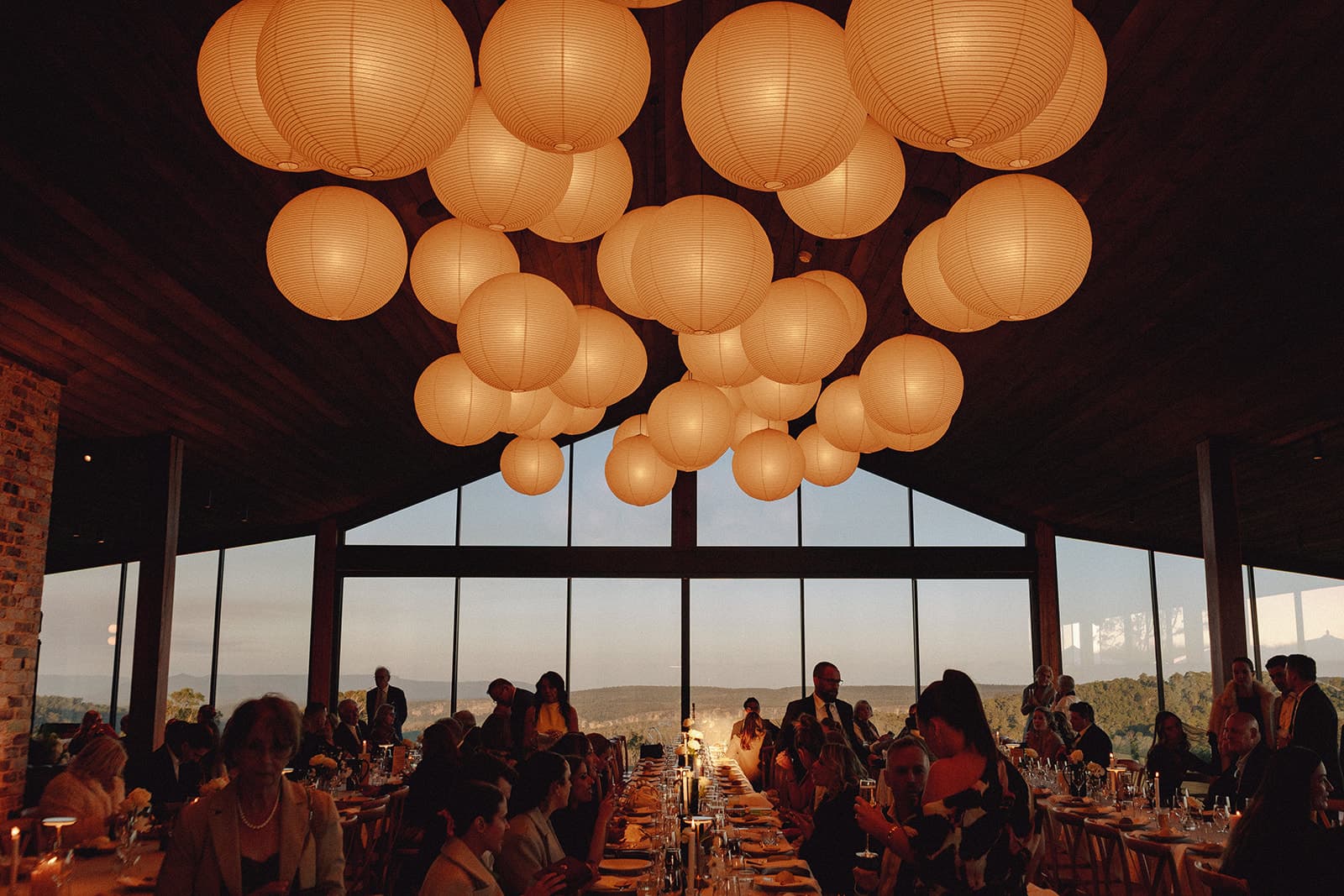Paper lanterns glowing above the long tables inside the Pavilion at dusk, guests seated the length of the room and the gorge still light through the glass gable